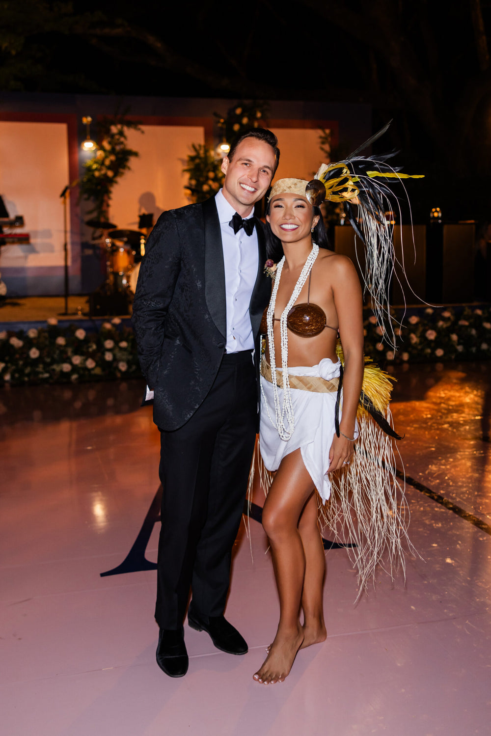 Bride and groom pose at a wedding reception, with the bride wearing a Tahitian-inspired outfit and feathered headdress