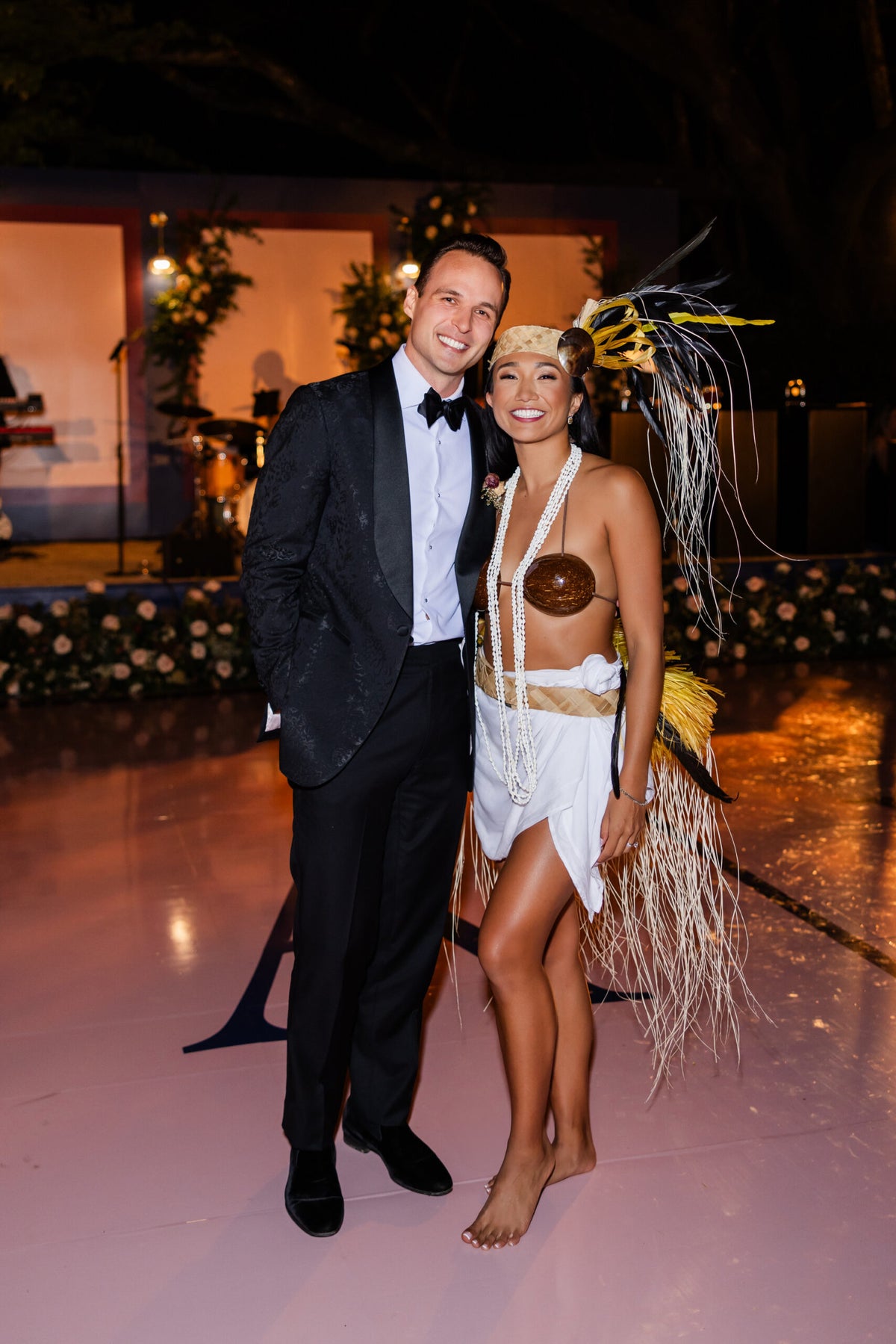 Bride and groom pose at a wedding reception, with the bride wearing a Tahitian-inspired outfit and feathered headdress