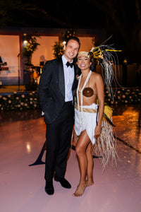 Bride and groom pose at a wedding reception, with the bride wearing a Tahitian-inspired outfit and feathered headdress