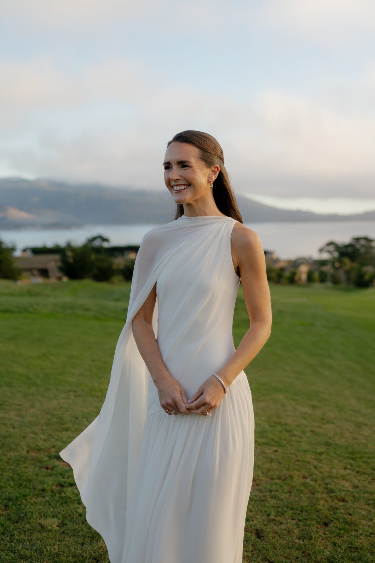 Emmie McKiernan smiles in a white wedding dress on a lawn with water and hills behind her