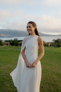 Emmie McKiernan smiles in a white wedding dress on a lawn with water and hills behind her