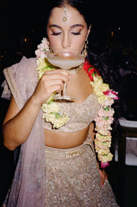 Bride holding a cocktail while wearing an ornate silver embellished lehenga and floral garlands in a dimly lit setting