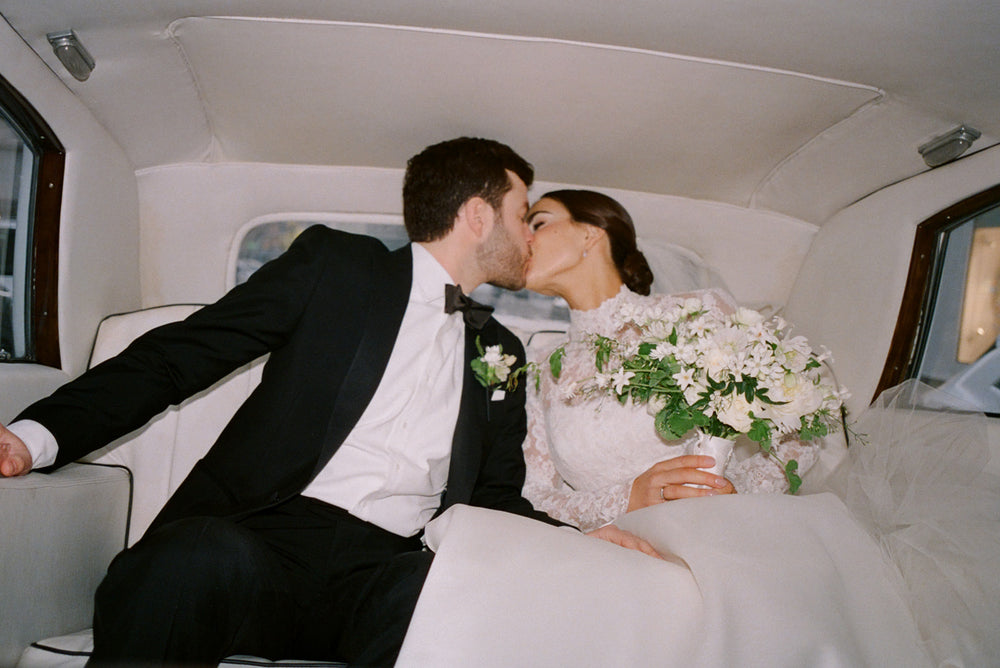 Bride and groom kissing in the back of a car, with the bride holding a white bouquet