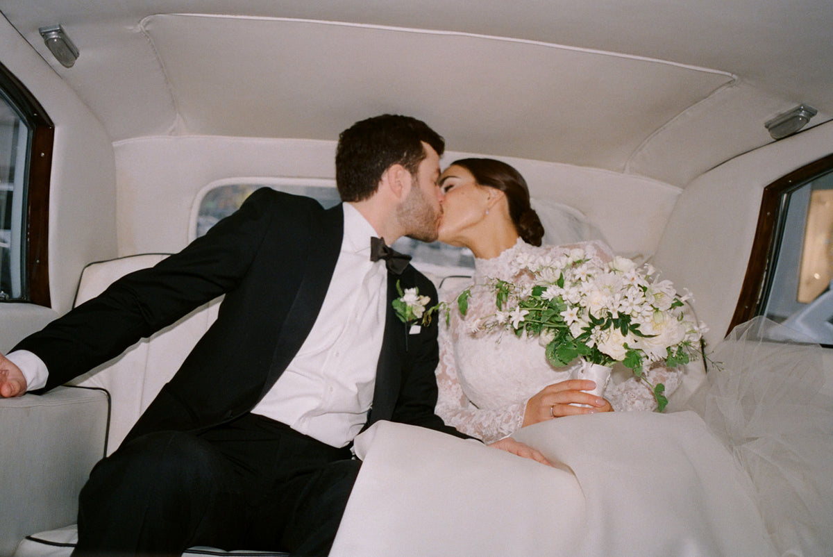 Bride and groom kissing in the back of a car, with the bride holding a white bouquet