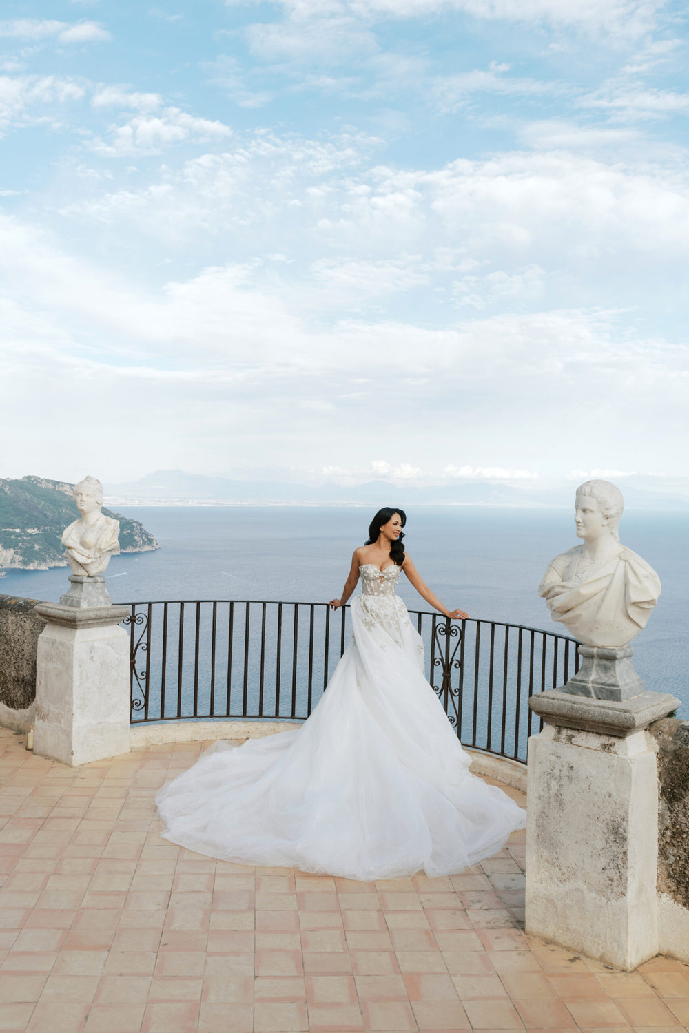 Bride in a white wedding gown poses between marble statues overlooking the sea at Villa Cimbrone