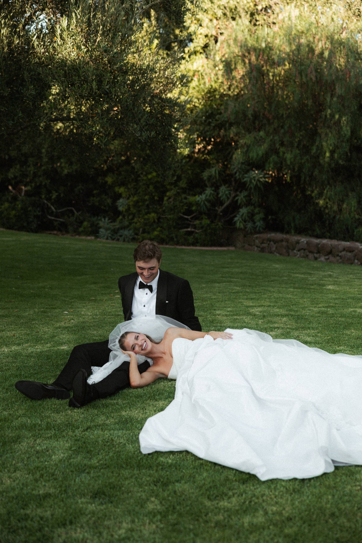 Newlyweds Lou Martines and Mac Kaufman relax on a lawn, with her white wedding dress spread across the grass