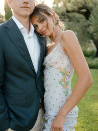 Maggie Varvel and Derek Vincent pose together outdoors in Provence, she wears a white floral dress and he wears a dark suit