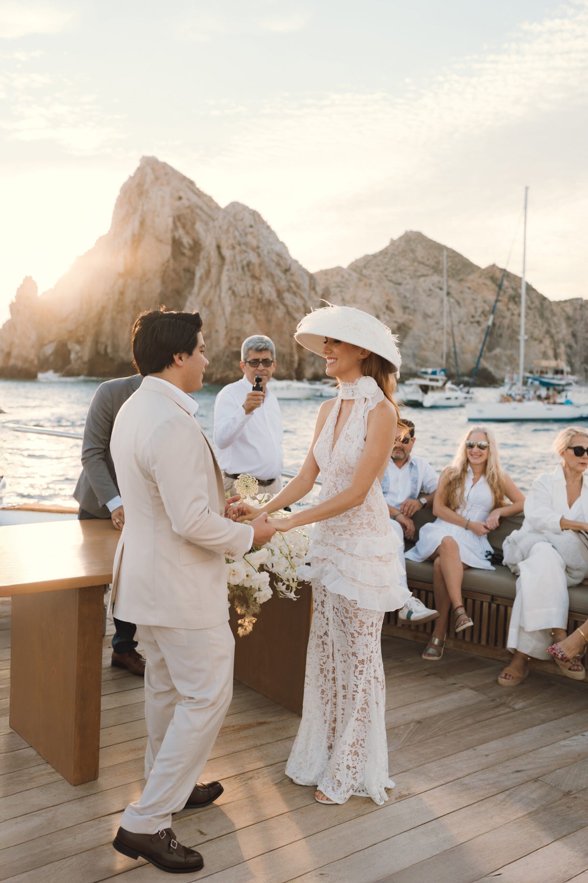 Bride and groom hold hands during a boat wedding ceremony in Baja, with guests and rocky coastline behind them