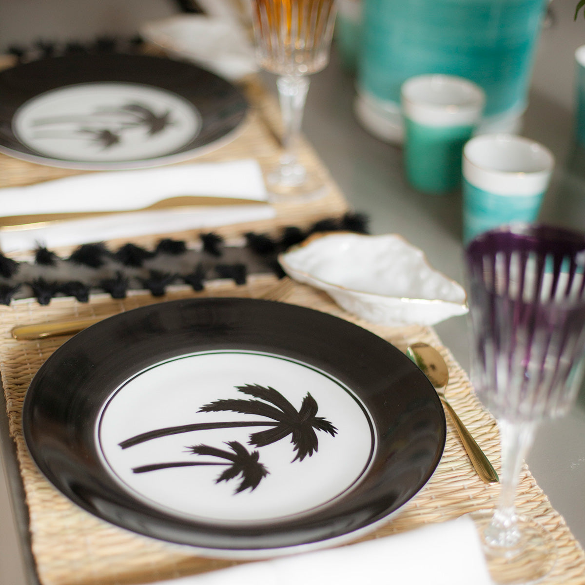 Angled view of Palm Beach charger plate showing black palm tree motif on white center and glossy black rim on woven placemat