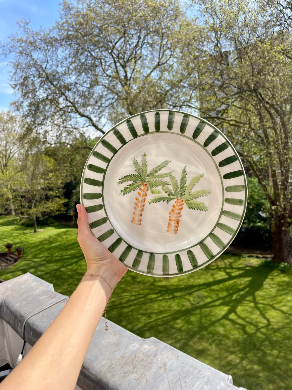 Front view of Marrakech palm tree ceramic plate with two hand-painted palms and green striped rim held against sunlit garden