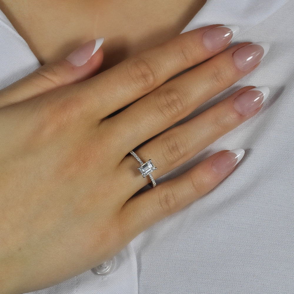Close-up of hand wearing Paradis emerald-cut diamond solitaire ring showcasing emerald-cut center and pavé band on white fabric background