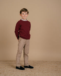Young boy wearing a maroon sweater and beige pants standing against a beige background