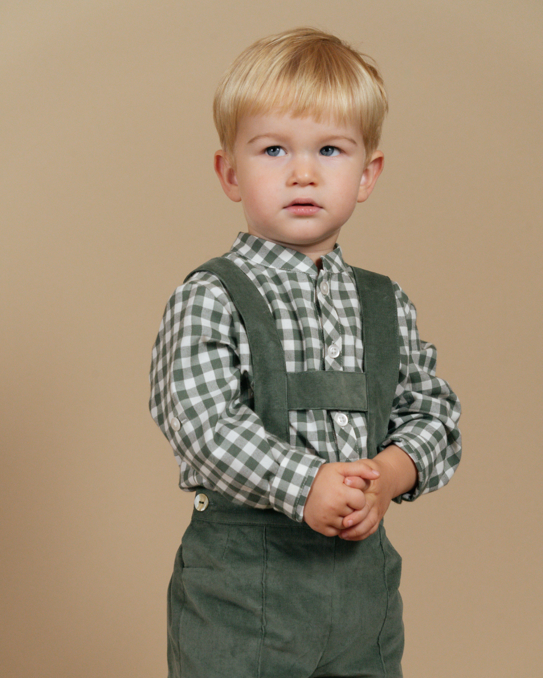 Child wearing a checkered shirt and green suspenders against a beige background
