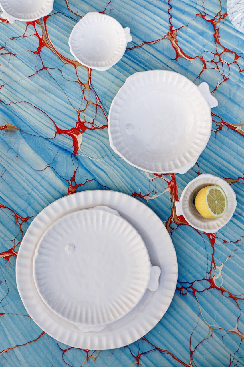 Top down view of Pesce Serena round platter showcasing scalloped rim and fish tail handle in white stoneware on blue marbled background