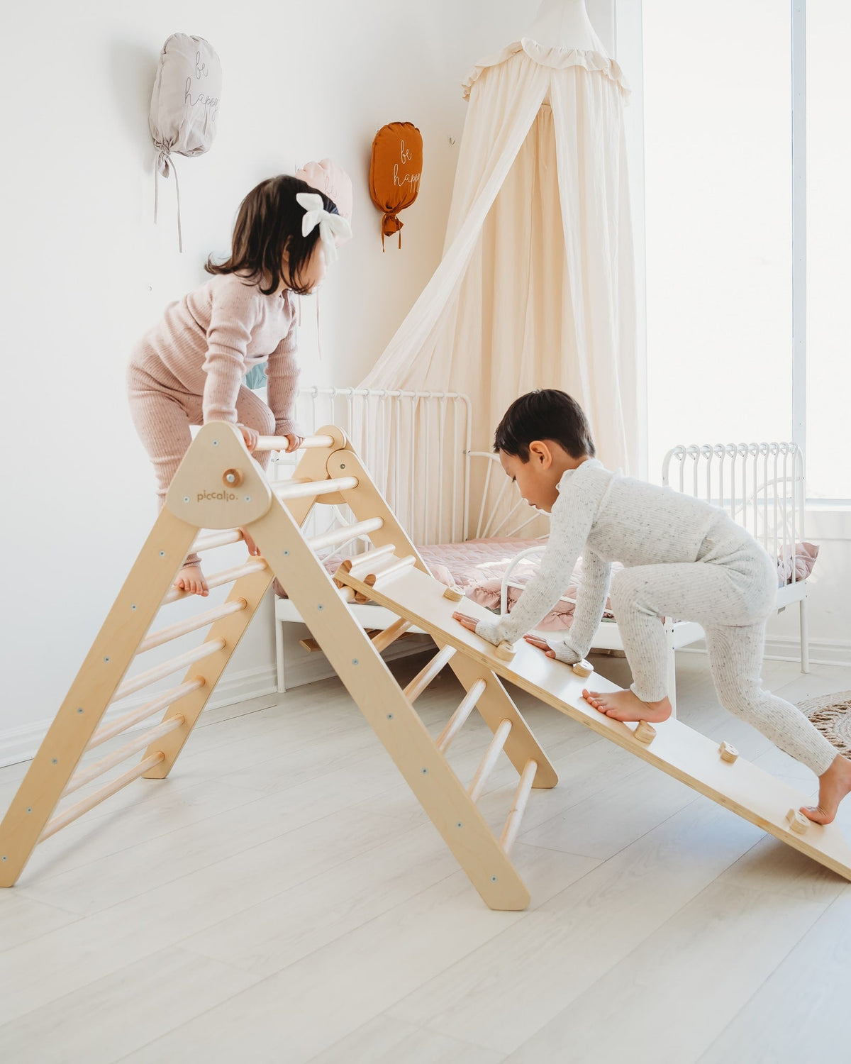 Angled view of Piccalio wooden Pikler triangle with climbing ramp and ladder rungs, two toddlers playing in a bright nursery