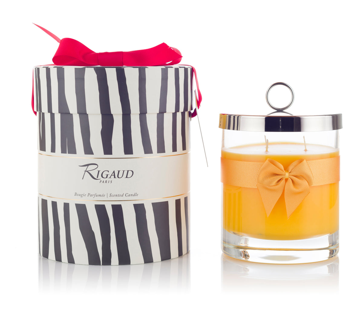 Front view of Rigaud Tournesol scented candle in clear glass with orange wax, bow and silver lid beside zebra-striped gift box with pink ribbon