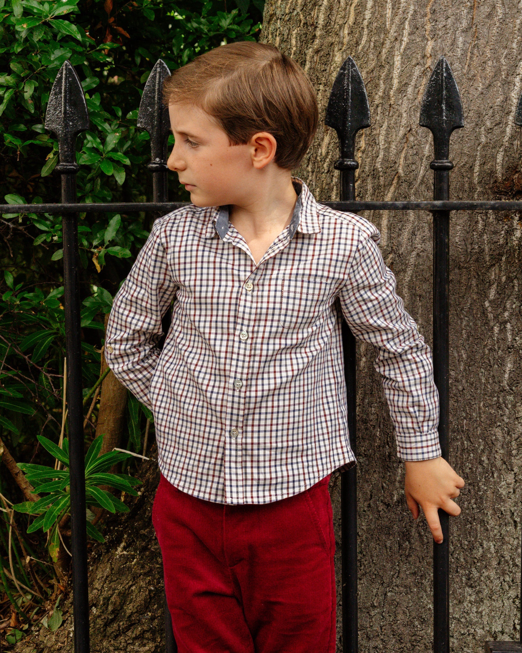 Young boy in a checkered shirt and red pants standing against a black metal fence.