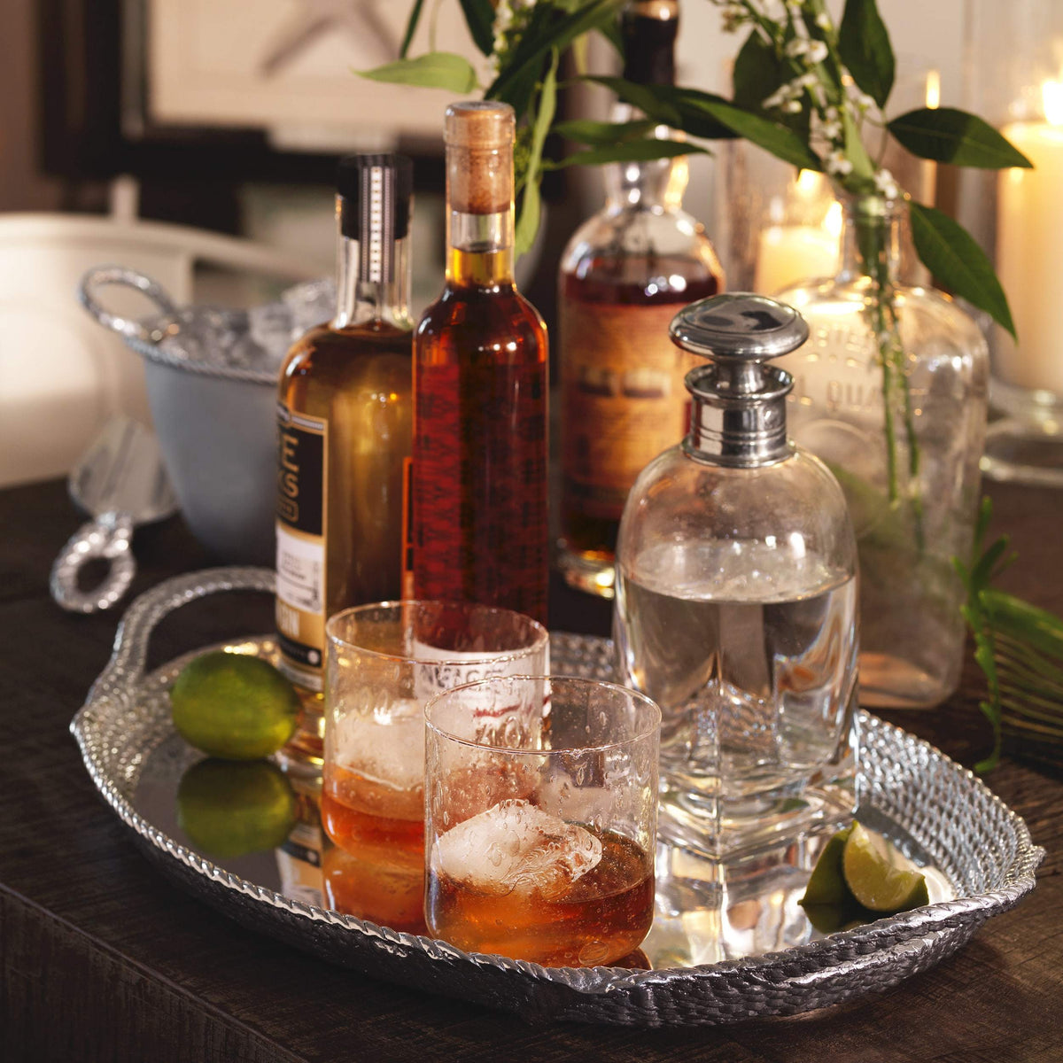 Rope handled ice scoop on a hammered silver tray with two whiskey glasses and bottles on a dark wood bar