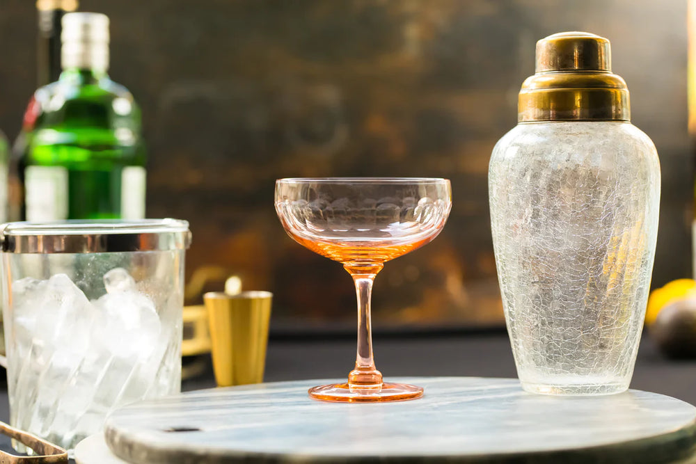 Front view of rose coupe cocktail glass showing engraved lens band and stem on tabletop with shaker and ice bucket
