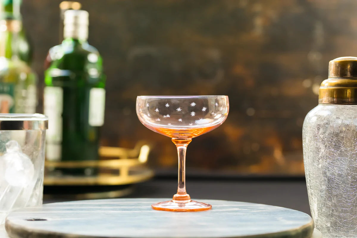 Front view of rose-colored coupe cocktail glass showcasing engraved star pattern with bar setting