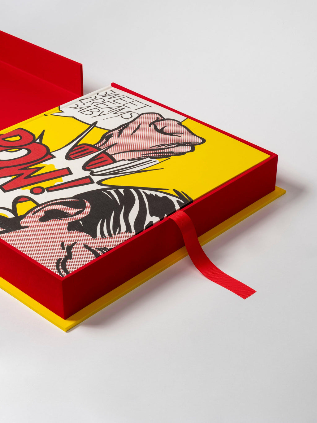 The Impossible Collection of Roy Lichtenstein by Assouline: Top-down view of the book flat in its open red presentation box. The book cover shows SWEET DREAMS, BABY! in a white comic speech bubble on yellow, POW! in large red letters below, and a dark-haired figure rendered in Ben-Day dots. A red ribbon pull is visible along the spine.