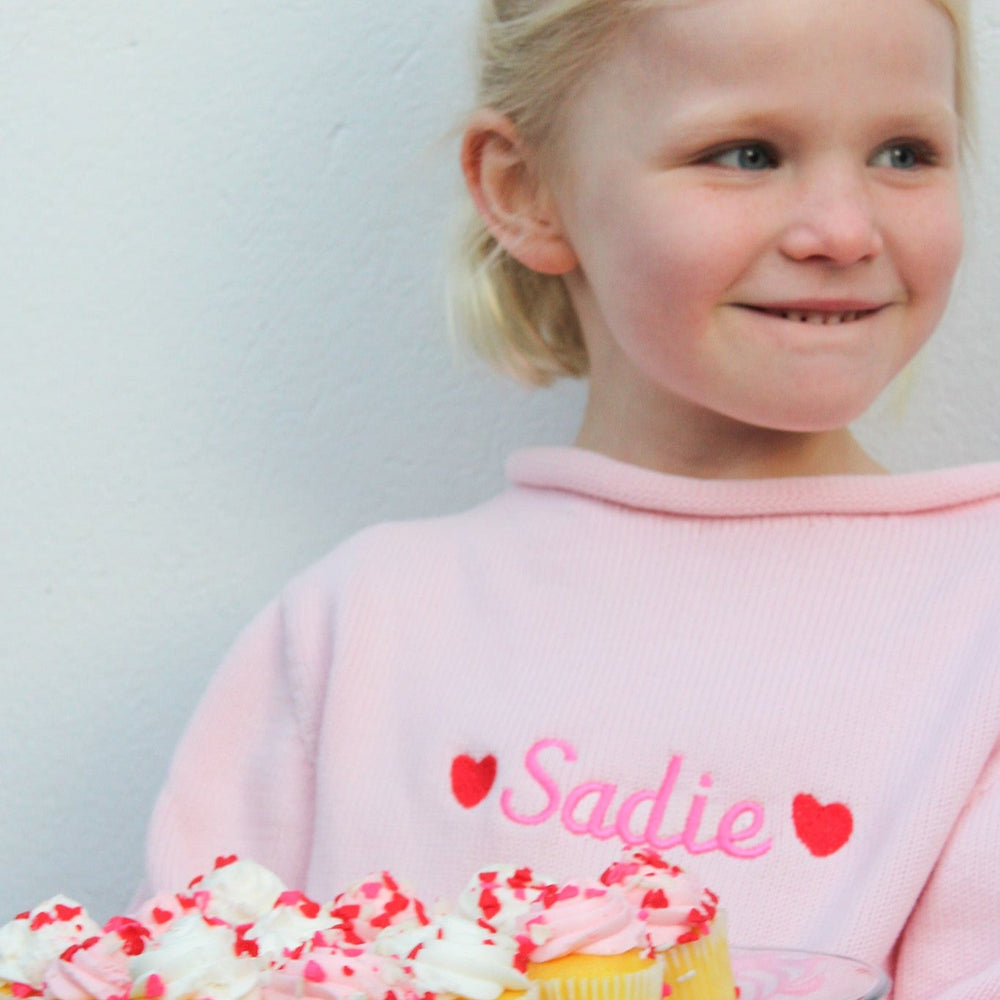 Front view of pink children’s rollneck sweater on child model showing embroidered name Sadie and red hearts against light wall