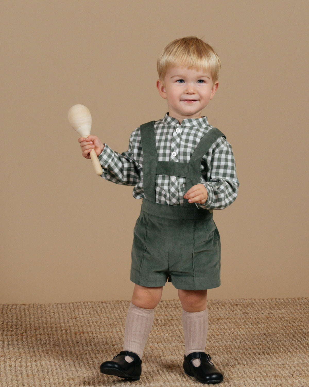Child wearing green overalls and a checkered shirt, holding a wooden rattle against a beige background
