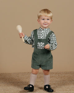 Child wearing green overalls and a checkered shirt, holding a wooden rattle against a beige background