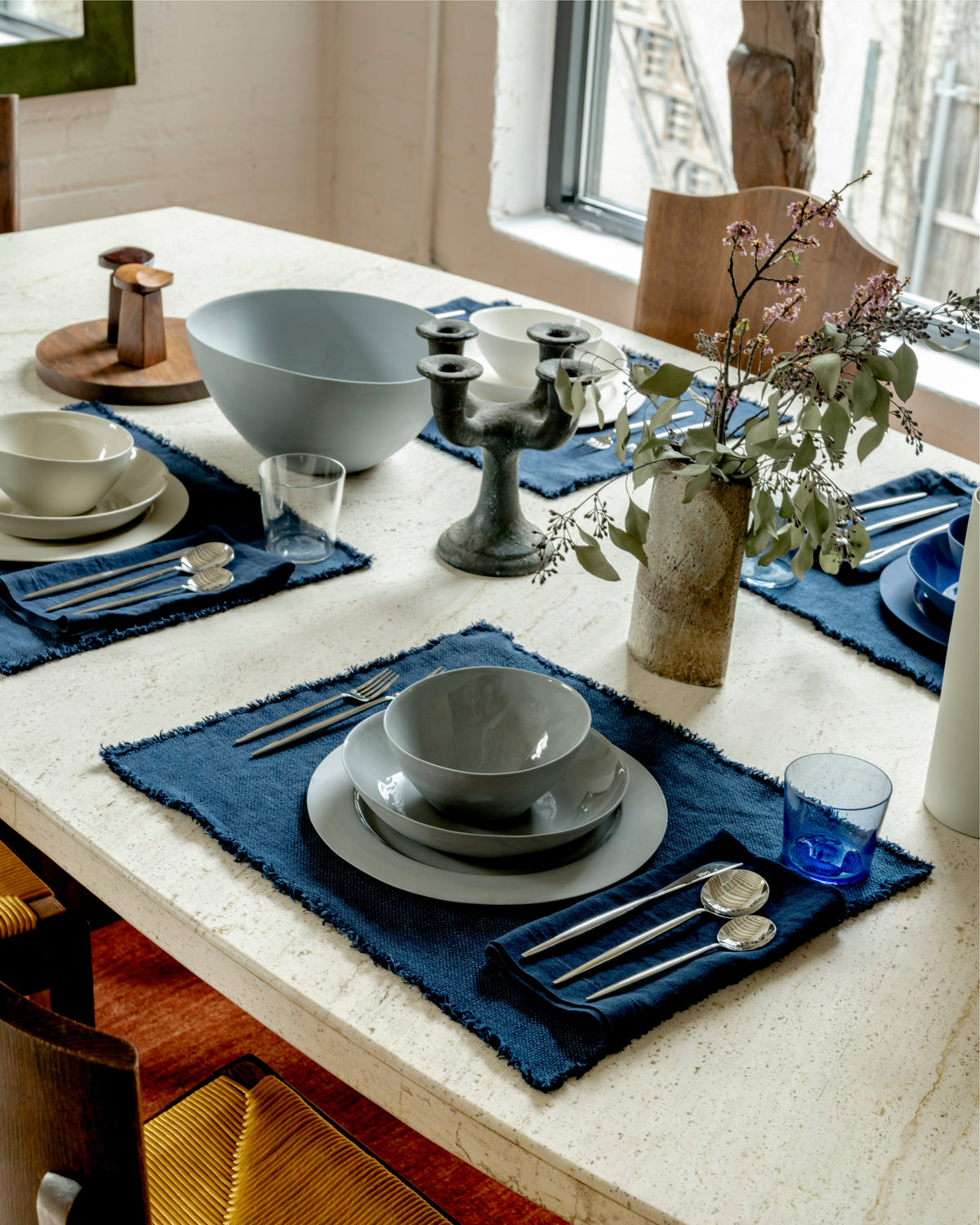 Blue linen placemats arranged with dinnerware on a dining table beside flowering branches
