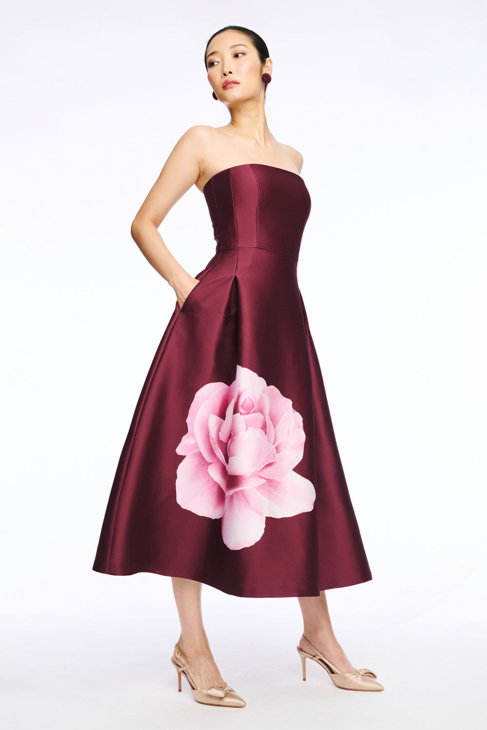 Three-quarter view of Shay Dress in Bordeaux with strapless bodice, A-line skirt with large pink flower print and pocket on white
