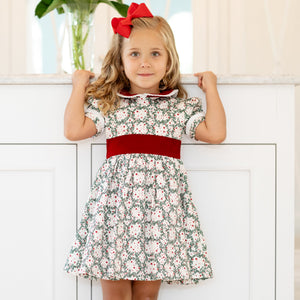Girls’ holiday dress in Henry Duvall Regent Wreaths red and green Christmas print, featuring a red velvet Peter Pan collar and sash, white pleated trim, puff sleeves, and a full gathered skirt.