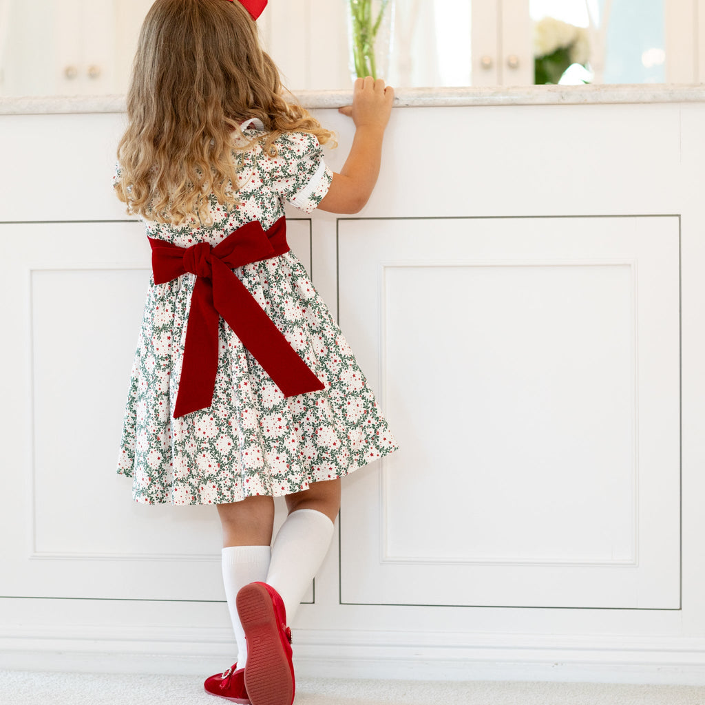 Girls’ holiday dress in Henry Duvall Regent Wreaths red and green Christmas print, featuring a red velvet Peter Pan collar and sash, white pleated trim, puff sleeves, and a full gathered skirt.