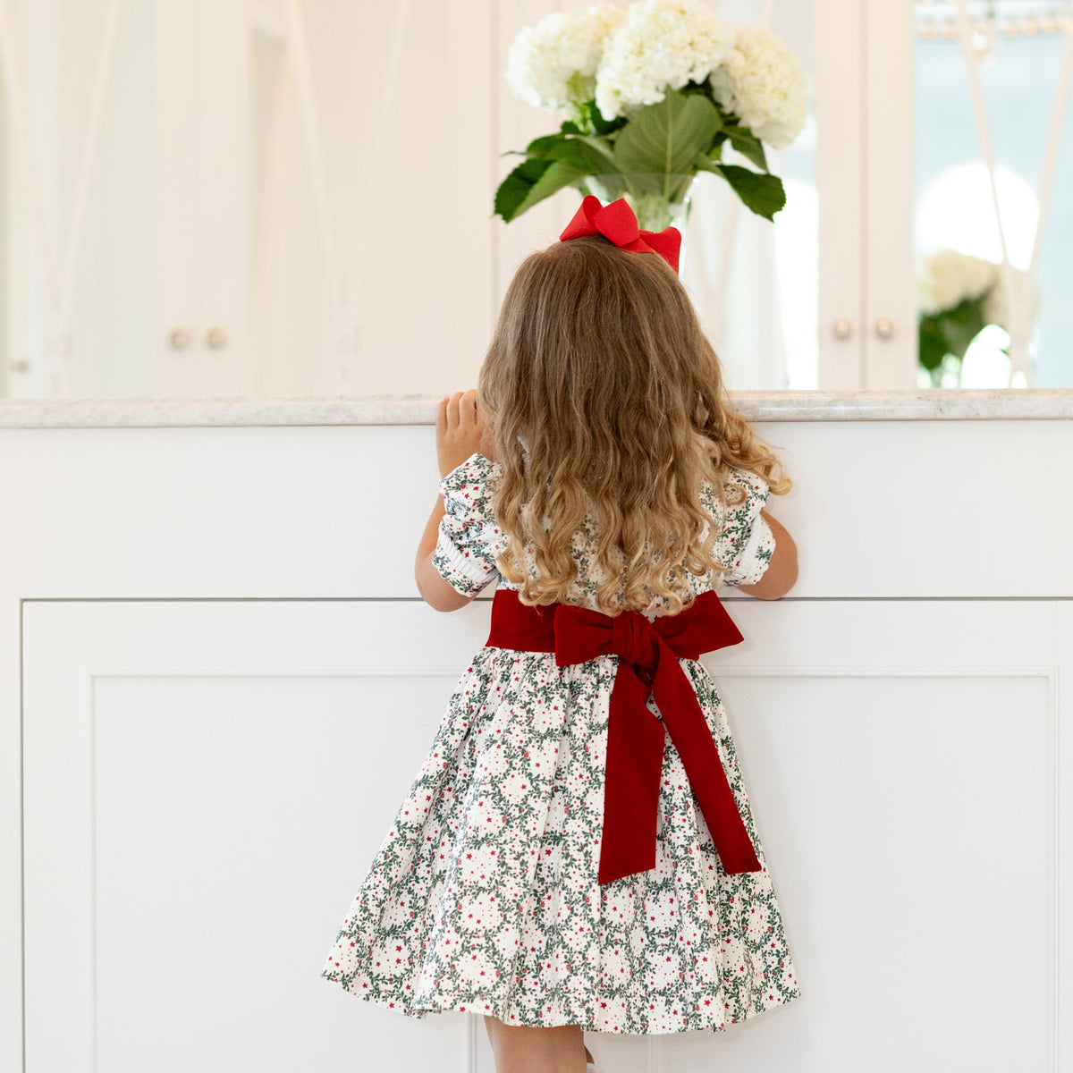 Back view of Short Sleeve Grace Dress in Regent Wreaths with red velvet sash and full gathered skirt against a white vanity
