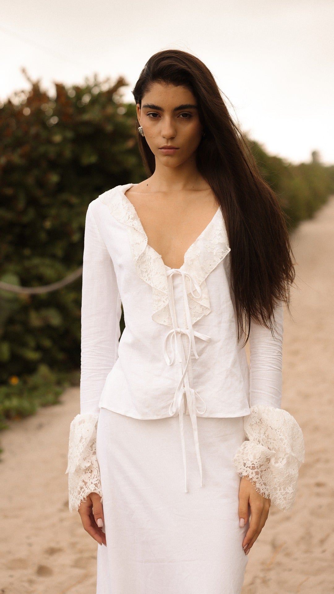 Model walking on a sandy path wearing a white lace-trimmed tie-front top and matching white skirt from Sister NY.