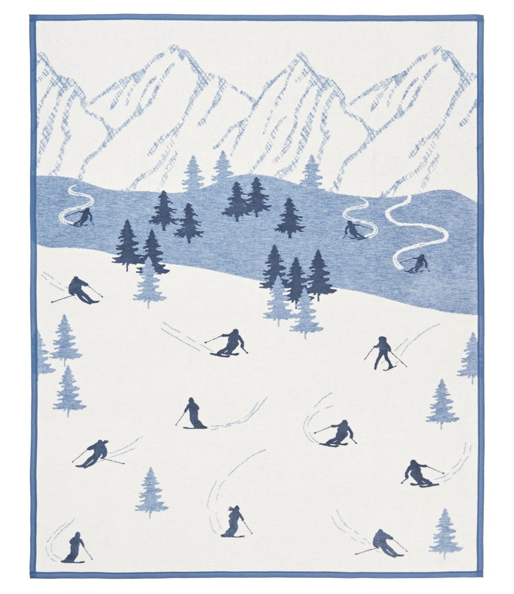 Cream and blue blanket featuring skiers on snowy slopes with evergreen trees and snow-capped mountains.