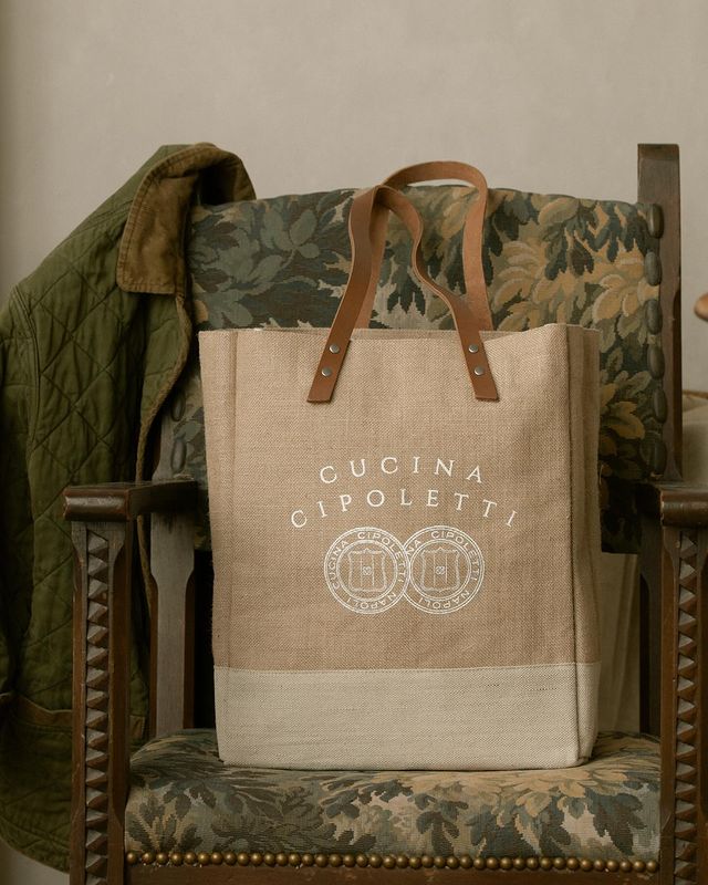 Front view of Cucina Cipoletti wine tote showcasing printed logo, leather straps, and two-tone canvas on a patterned chair