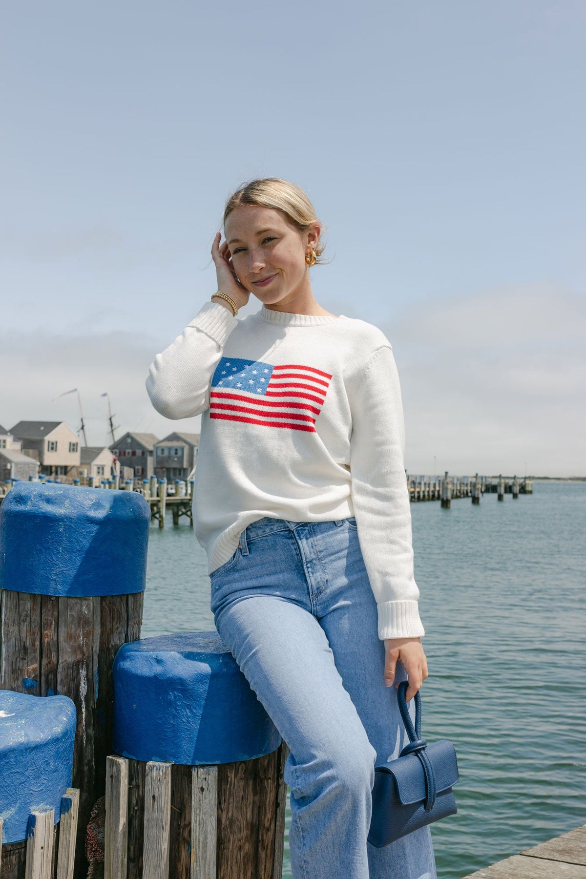 Model wearing an ivory American flag crewneck sweater posing on a waterfront dock