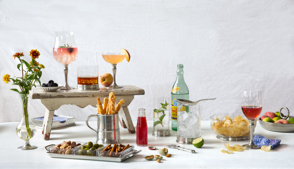Summer beverage spread with wine and cocktails, ice bucket, chips and fruit on a white tablecloth