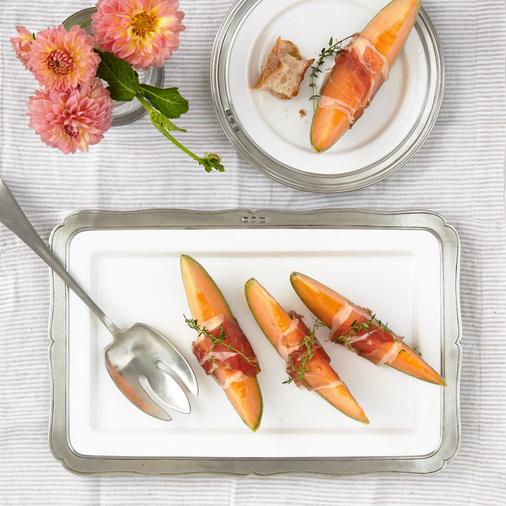 Top-down view of antique pewter serving fork with three-pronged head and matte luster on white tray with prosciutto-wrapped melon