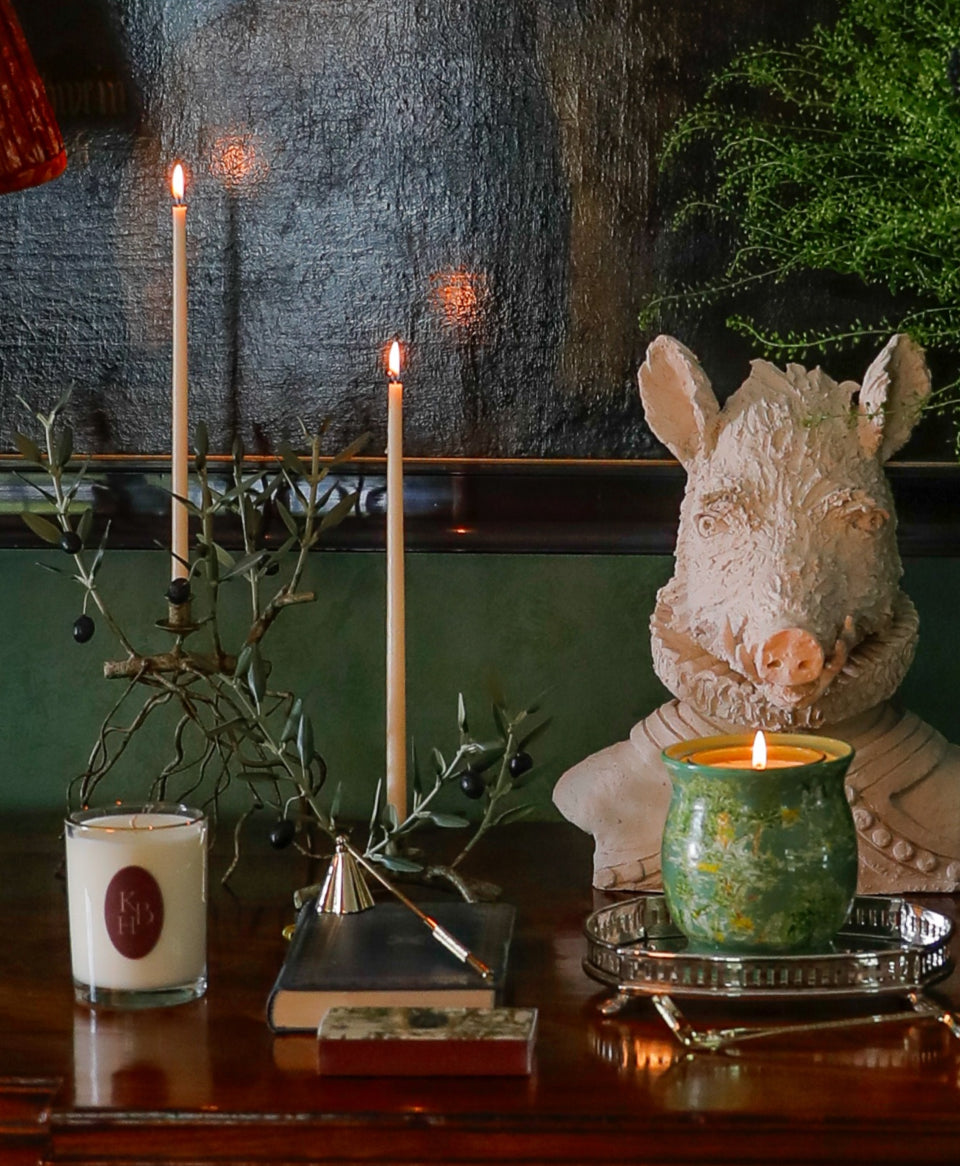 Signature Scent candle burning in green ceramic jar on silver tray beside decorative boar bust and lit tapers on wooden table