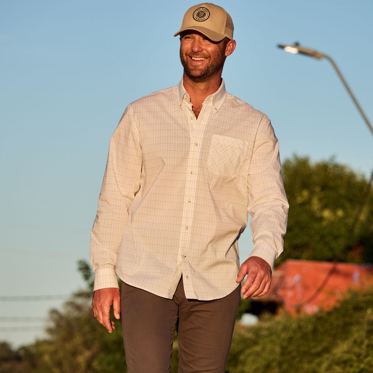 Man in a Powell Performance Shirt by Tom Beckbe and UPF 30+ beige cap, walking in a sunlit park with trees and streetlamp.