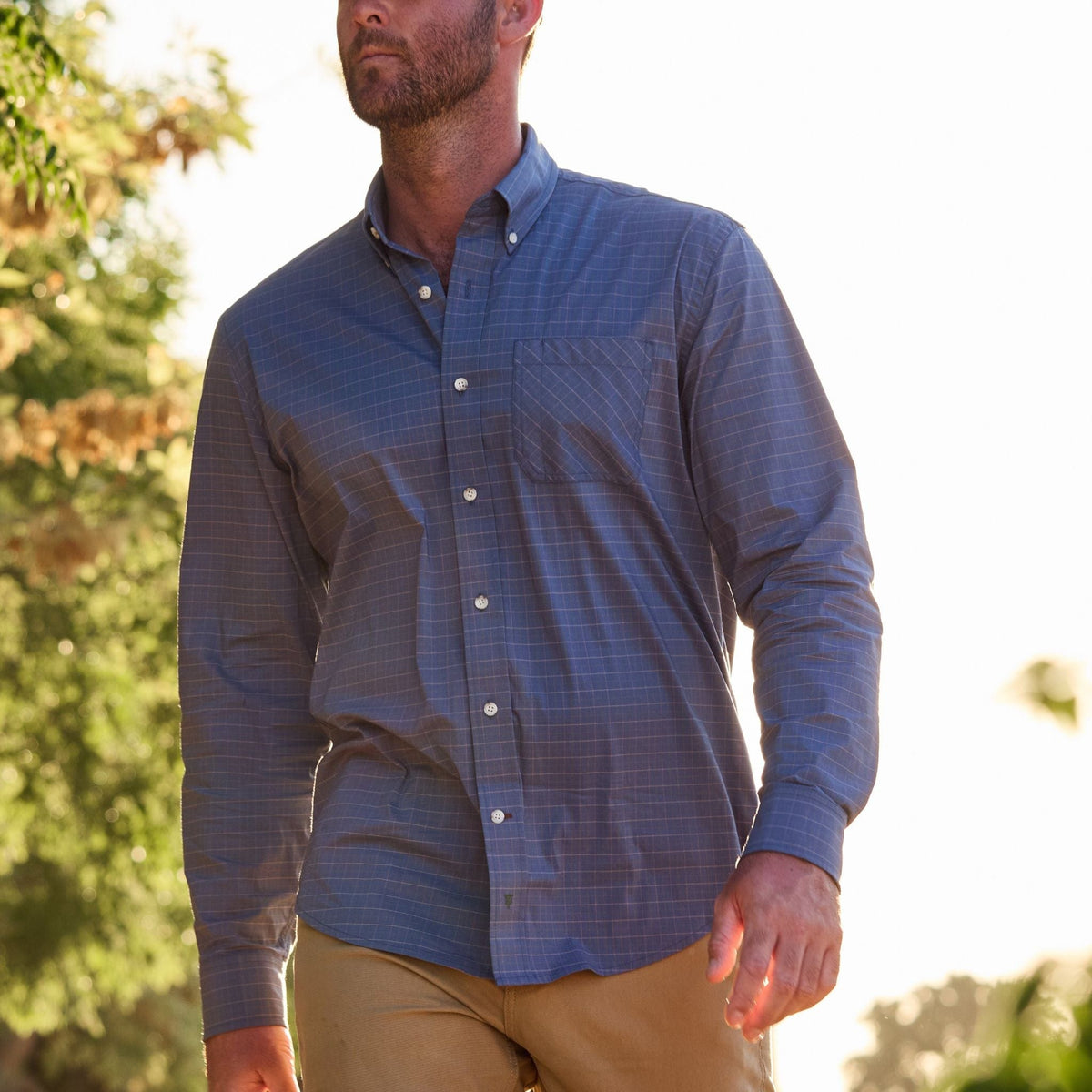 A man in a blue Tom Beckbe Powell Performance Shirt and khaki pants walks outdoors, enjoying the sunlight with trees behind.