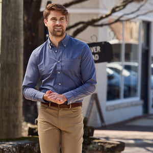 A man in a blue Tom Beckbe Powell Performance Shirt and khaki pants stands outside with hands clasped on a sunny urban day.