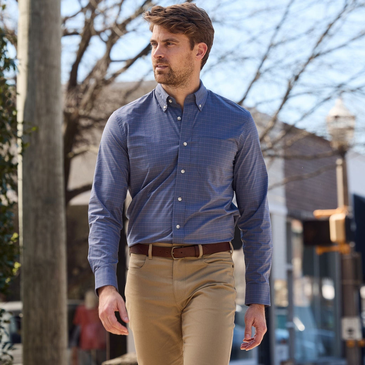 A man in a blue Tom Beckbe Powell Performance Shirt and khaki pants walks outdoors with trees and buildings behind him.