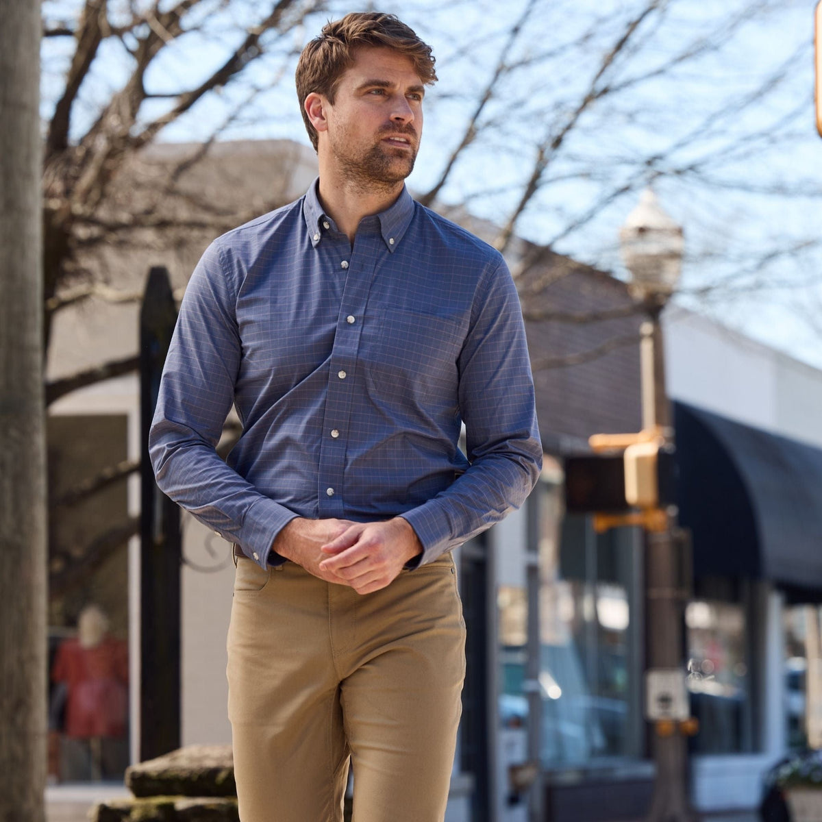 A man wearing a blue Tom Beckbe Powell Performance Shirt and khakis strolls on a sunny urban street with trees and shops.