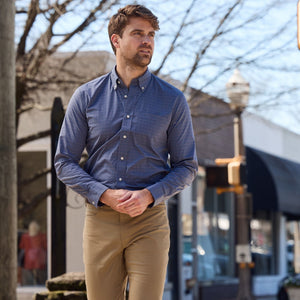 A man wearing a blue Tom Beckbe Powell Performance Shirt and khakis strolls on a sunny urban street with trees and shops.