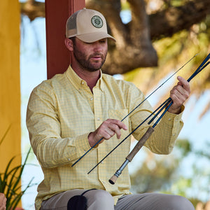 Man in a beige cap and Tom Beckbes Powell Performance Shirt assembling a fishing rod under a tree.