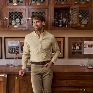 A man in a yellow Powell Performance Shirt by Tom Beckbe stands in a wooden bar, surrounded by bottles and framed covers.