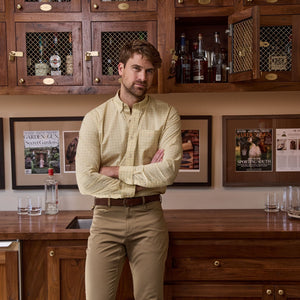 A man in a Tom Beckbe Powell Performance Shirt stands with crossed arms before a wooden bar with glass shelves.