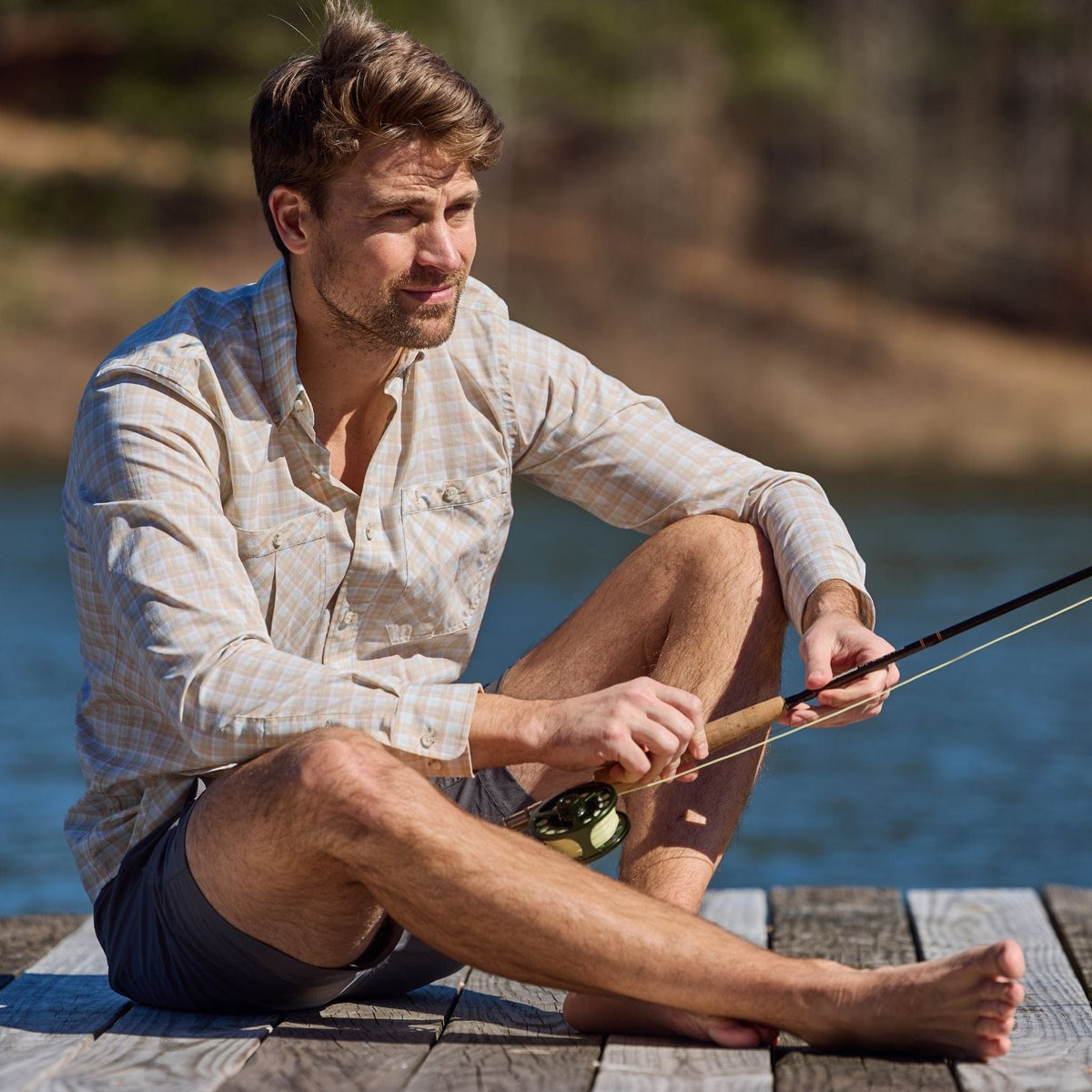 Man in a Tom Beckbe Tidewater Shirt is cross-legged on a dock, holding a fishing rod with water and trees behind him.